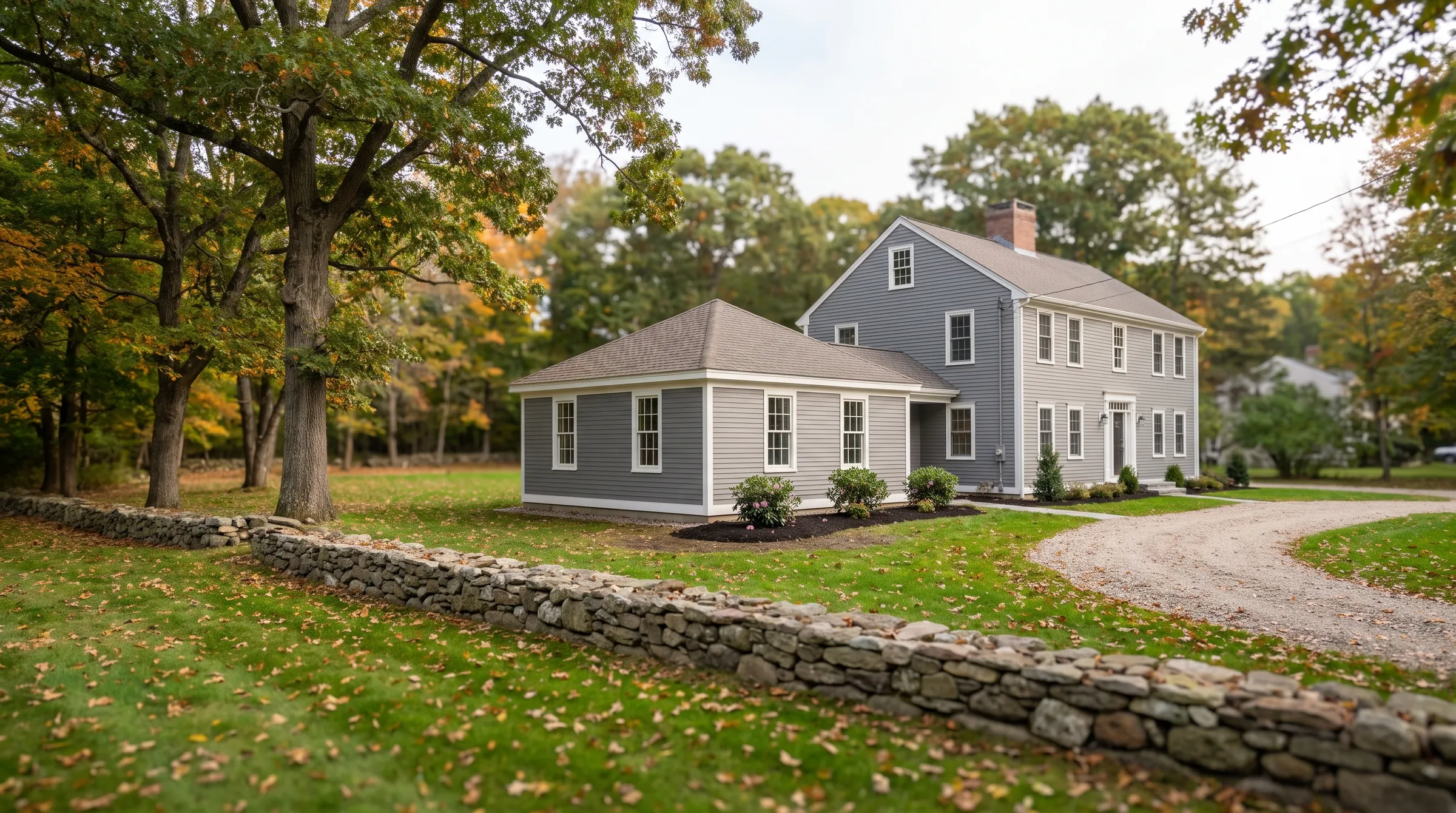 A gray center-chimney colonial on a wooded lot with a new single-story bedroom wing, a fieldstone wall and a gravel driveway