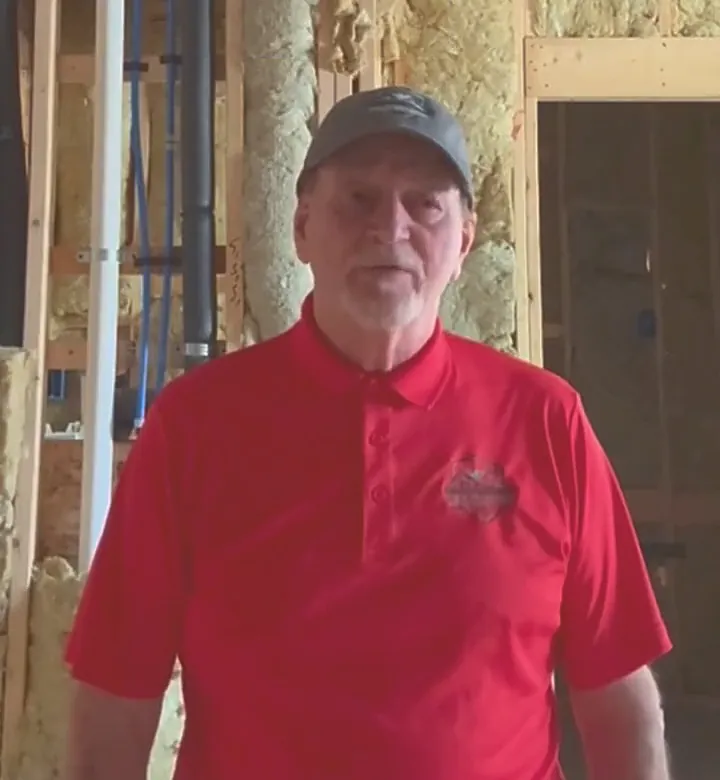 Stephen Selby in a red Renovation Defenders polo on a job site, open stud framing and insulation behind him